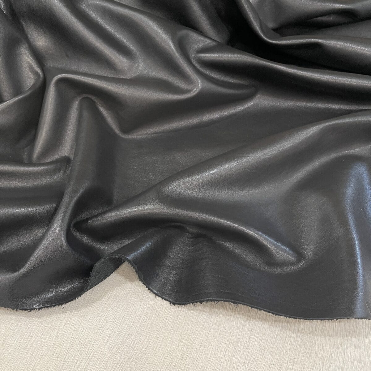 Dark gray, glossy leather fabric draped in soft folds and waves, showing texture and sheen on a light surface.