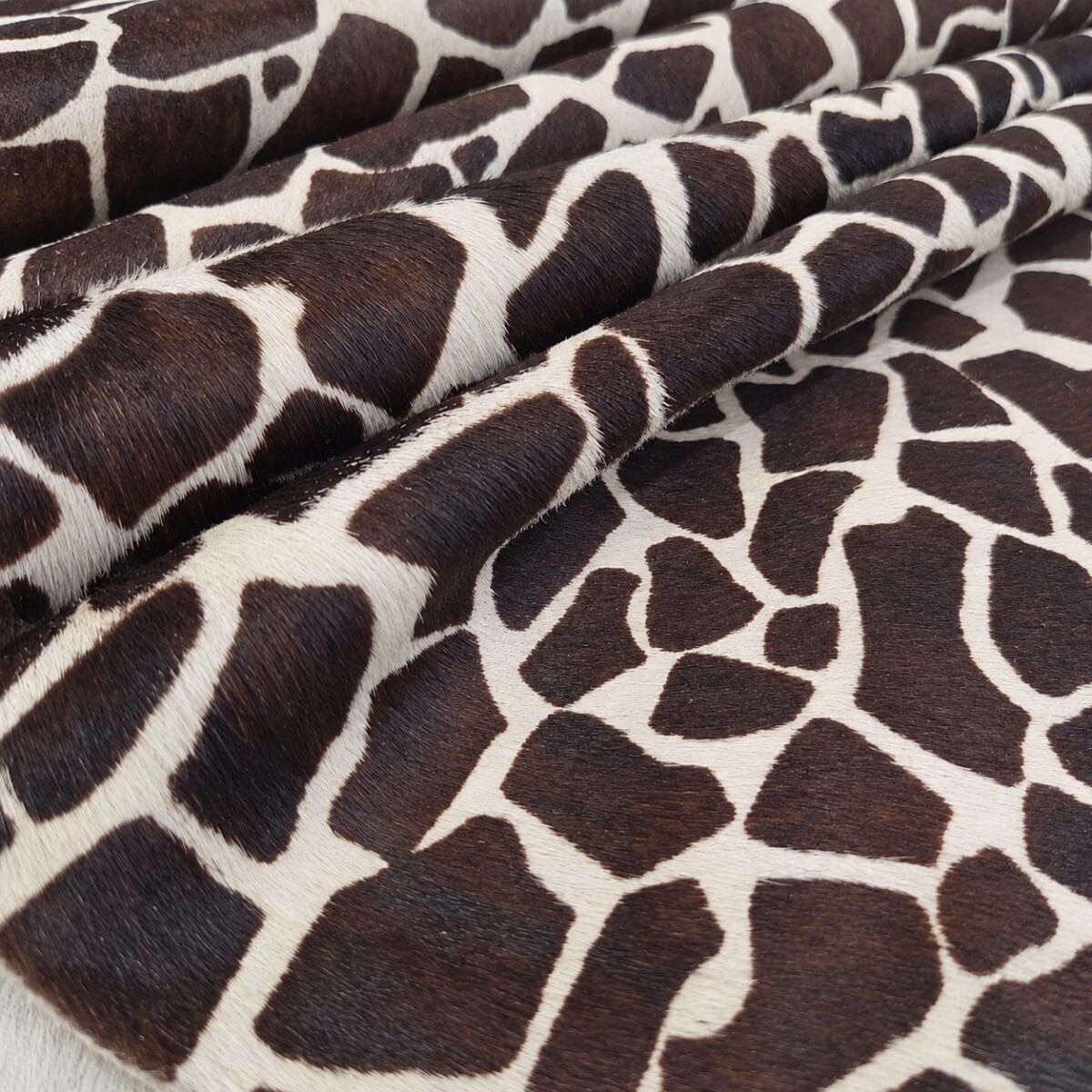 hair on cowhide giraffe print Close-up of rolled giraffe-patterned fabric with dark brown patches on a cream background, showing fuzzy texture and folds.
