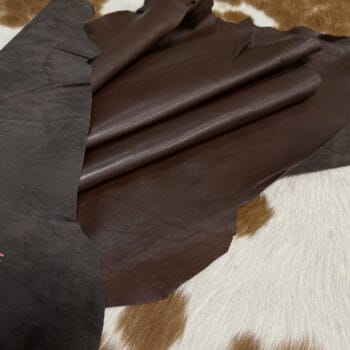 Dark brown leather swatches layered over a black suede sample, placed on a white-and-brown spotted fur background.
