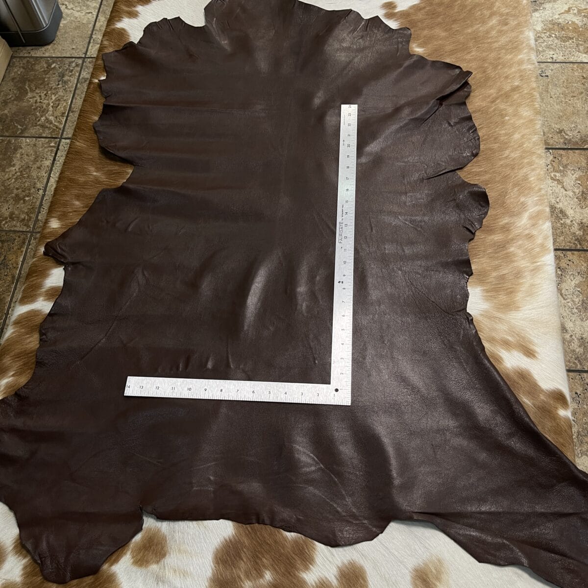 Dark brown leather hide laid flat on a light cowhide rug, with a metal ruler arranged in an L-shape across it for measurement