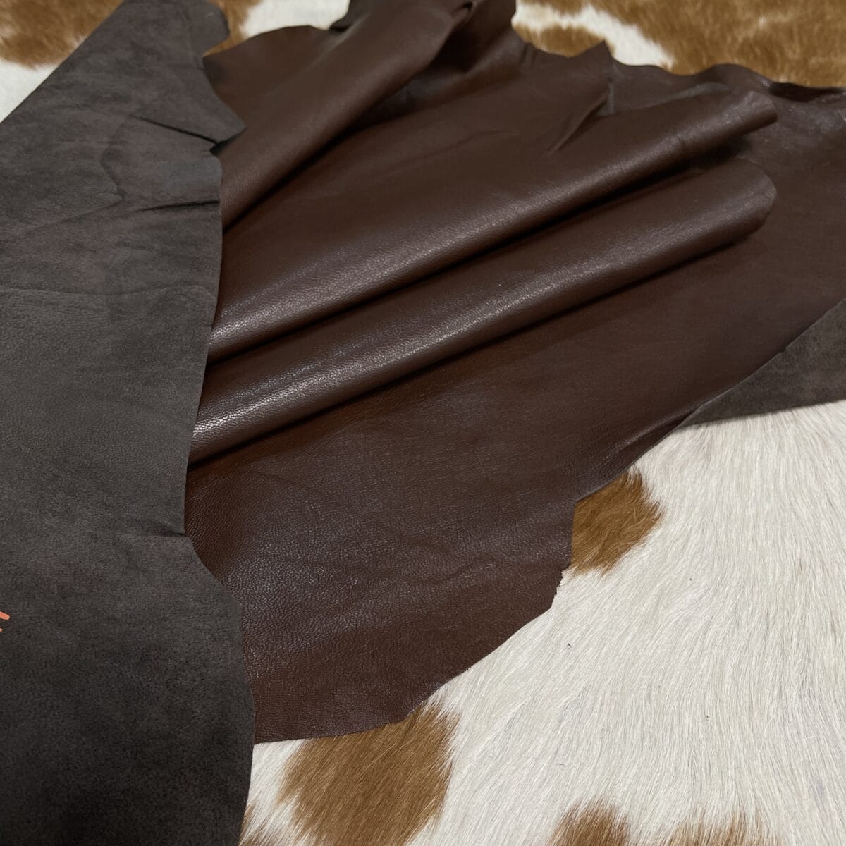 Dark brown leather swatches layered over a black suede sample, placed on a white-and-brown spotted fur background.