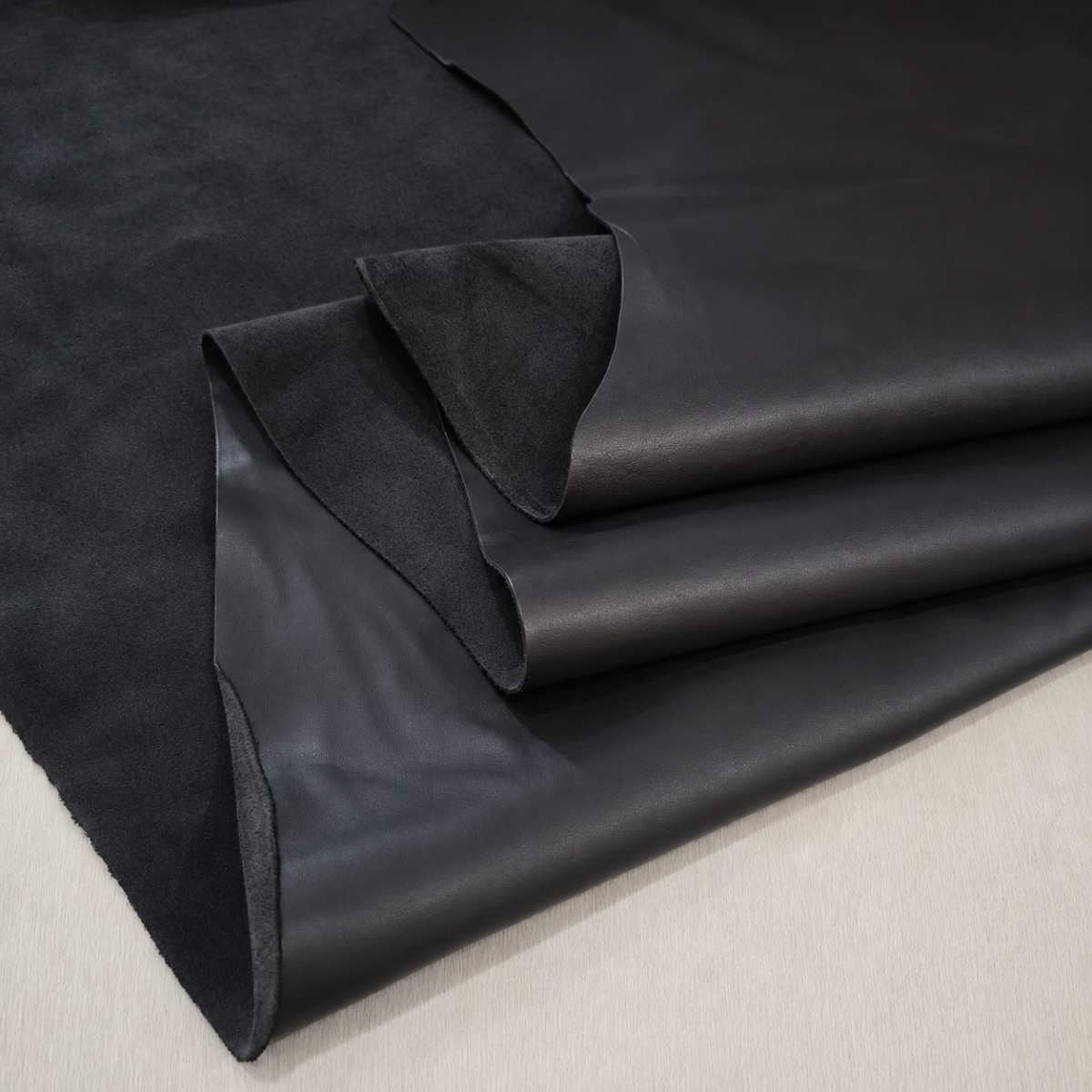 slblk skipper cow leather Stacks of black leather and suede hides folded and layered on a light surface, close-up view.